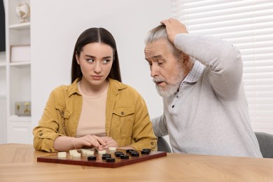 Playing checkers. Emotional senior man learning young woman at table in room