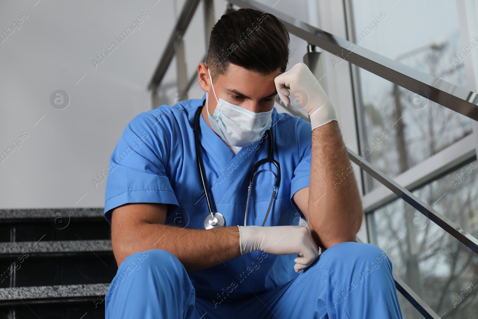 Photo of Sad doctor in facial mask on stairs indoors. Stress of health care workers during coronavirus pandemic