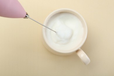 Whisking milk in cup with mini mixer (milk frother) on beige background, top view