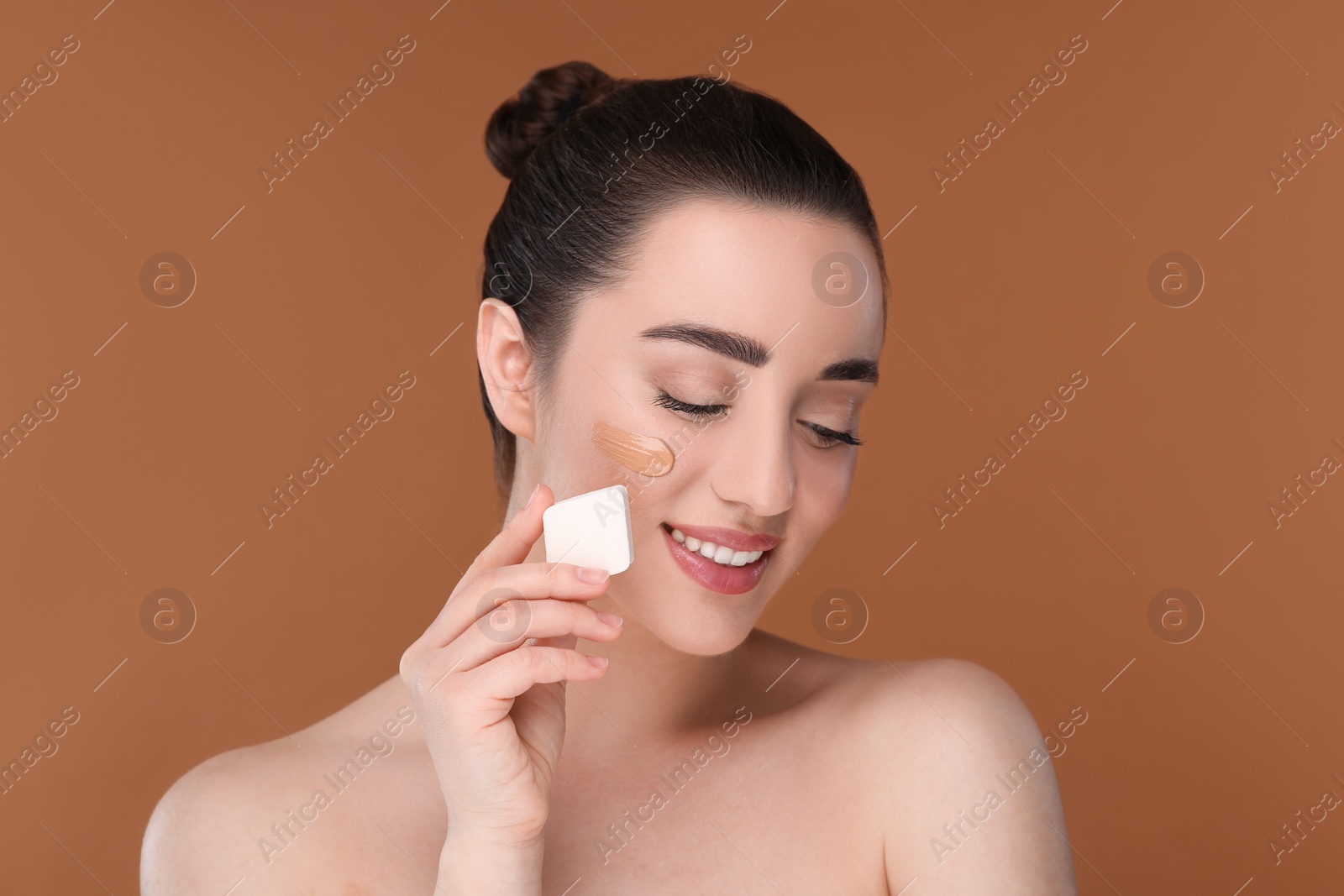 Photo of Woman applying foundation on face with makeup sponge against brown background