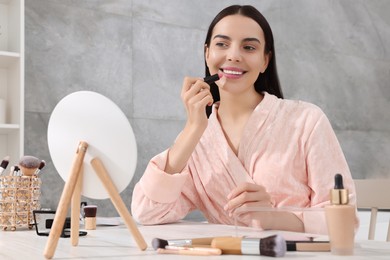 Photo of Beautiful young woman applying lipstick at dressing table indoors