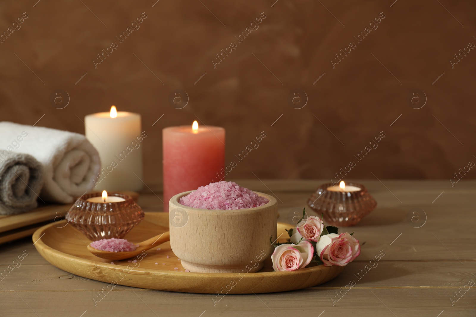 Photo of Bowl of pink sea salt, roses, burning candles, herbal massage bags and towels on wooden table