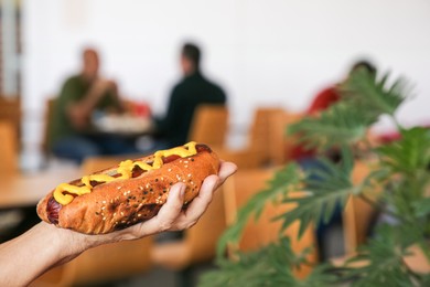 Photo of Woman holding fresh tasty hot dog with mustard in cafe, closeup. Space for text
