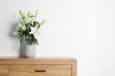 Beautiful bouquet of lily flowers in vase on chest of drawers near white wall, space for text