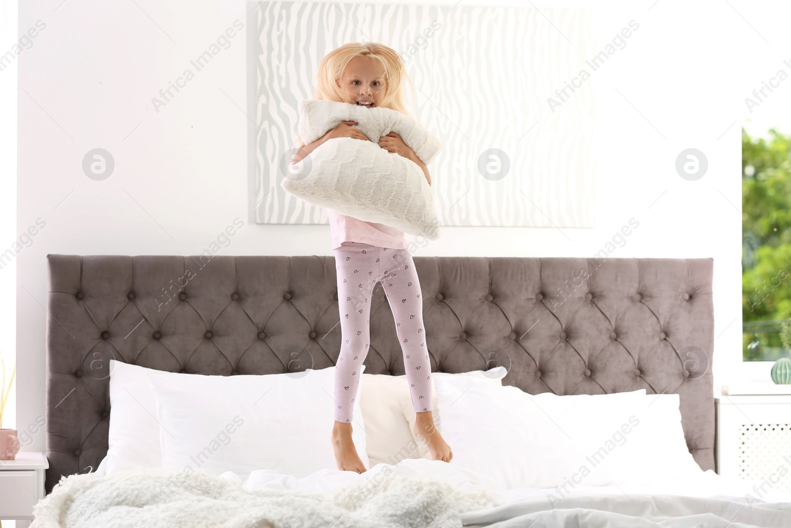 Photo of Cute little girl with pillow jumping on bed at home