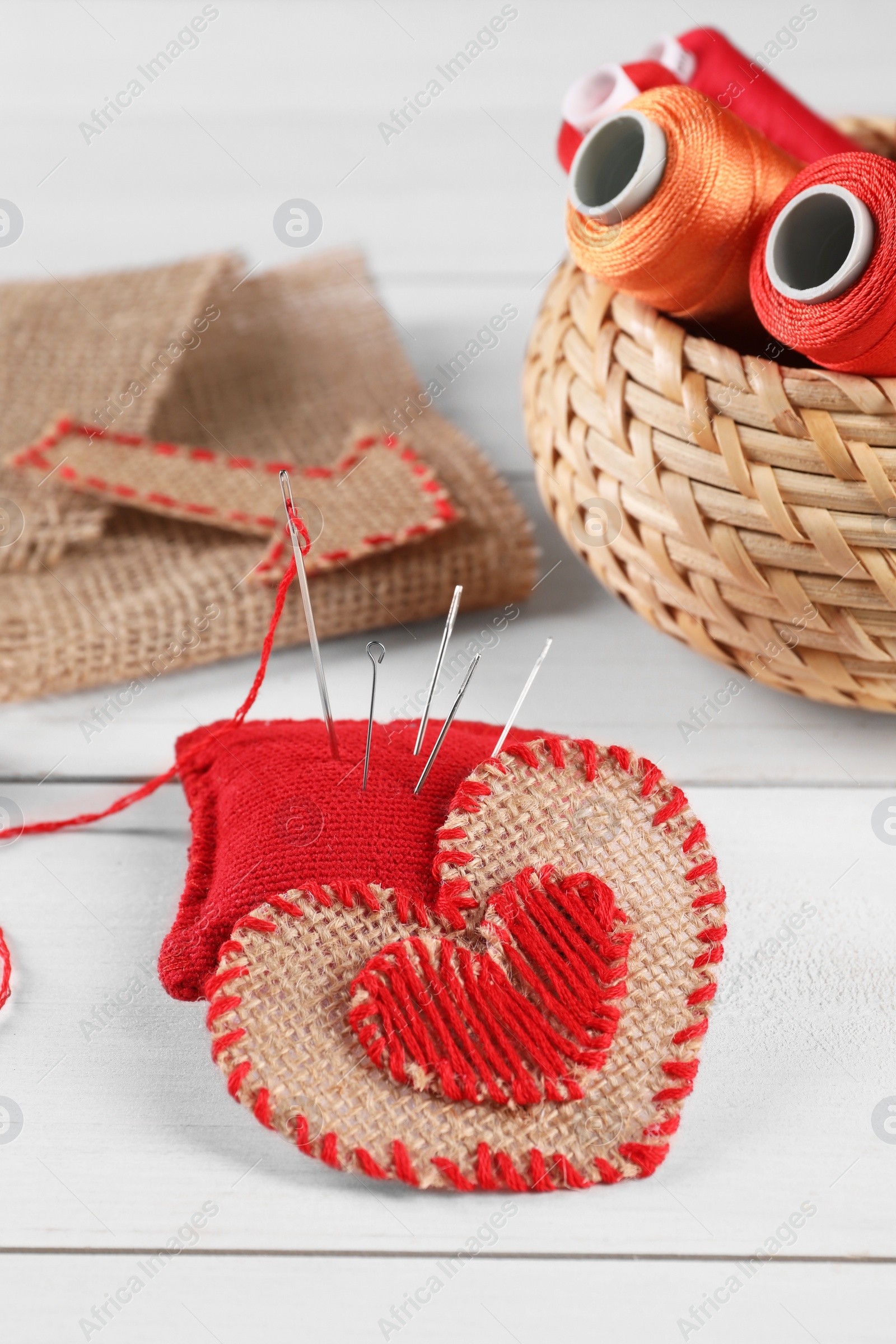 Photo of Heart of burlap fabric with red stitches, spools of threads and sewing tools on white wooden table