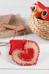 Photo of Heart of burlap fabric with red stitches, spools of threads and sewing tools on white wooden table