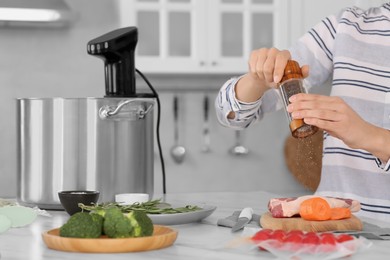 Photo of Woman grinding pepper onto meat near pot with sous vide cooker in kitchen, closeup. Thermal immersion circulator