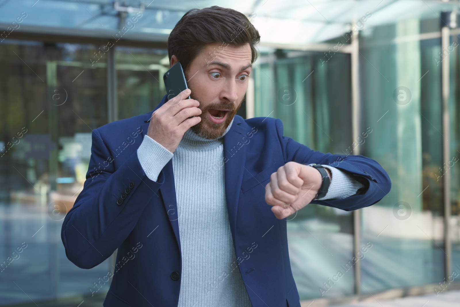 Photo of Emotional businessman talking on smartphone and looking at watch near building. Being late concept