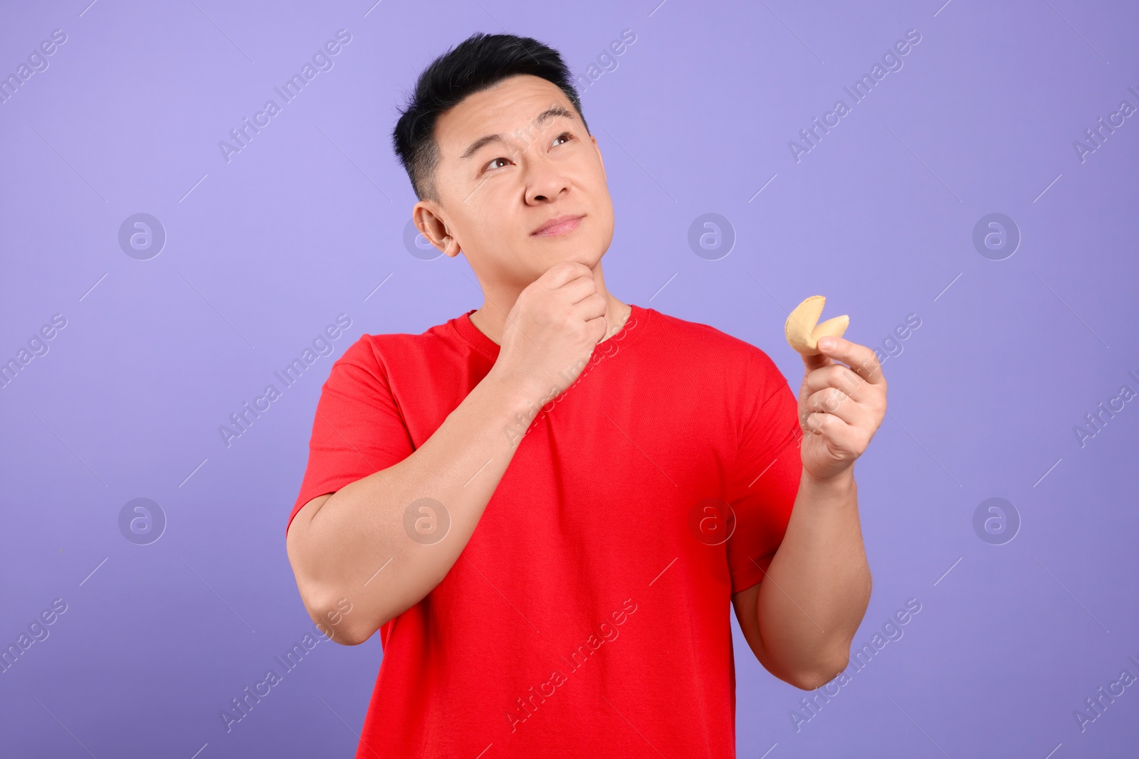 Photo of Asian man holding tasty fortune cookie with prediction on violet background