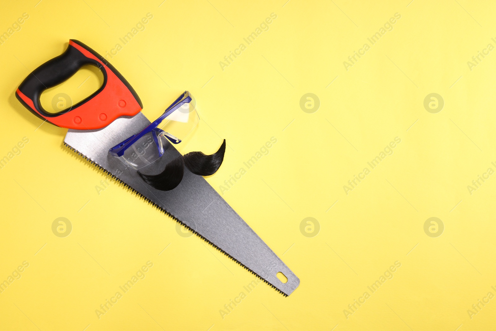 Photo of Man's face made of artificial mustache, safety glasses and hand saw on yellow background, top view. Space for text