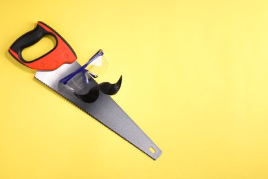 Man's face made of artificial mustache, safety glasses and hand saw on yellow background, top view. Space for text