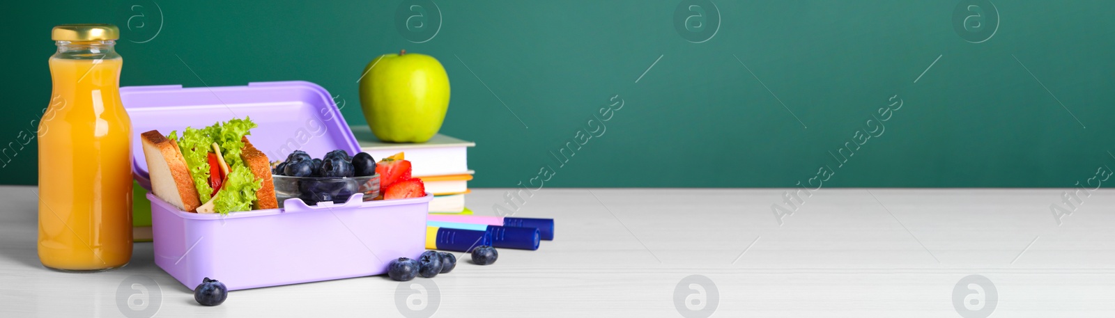 Image of School meal. Lunch box with healthy food and different stationery on white wooden table near green chalkboard, space for text. Banner design