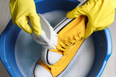 Photo of Woman with gloves and brush cleaning stylish sneakers in wash basin, top view