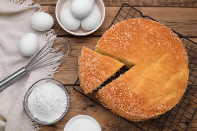 Tasty sponge cake, whisk and ingredients on wooden table, flat lay