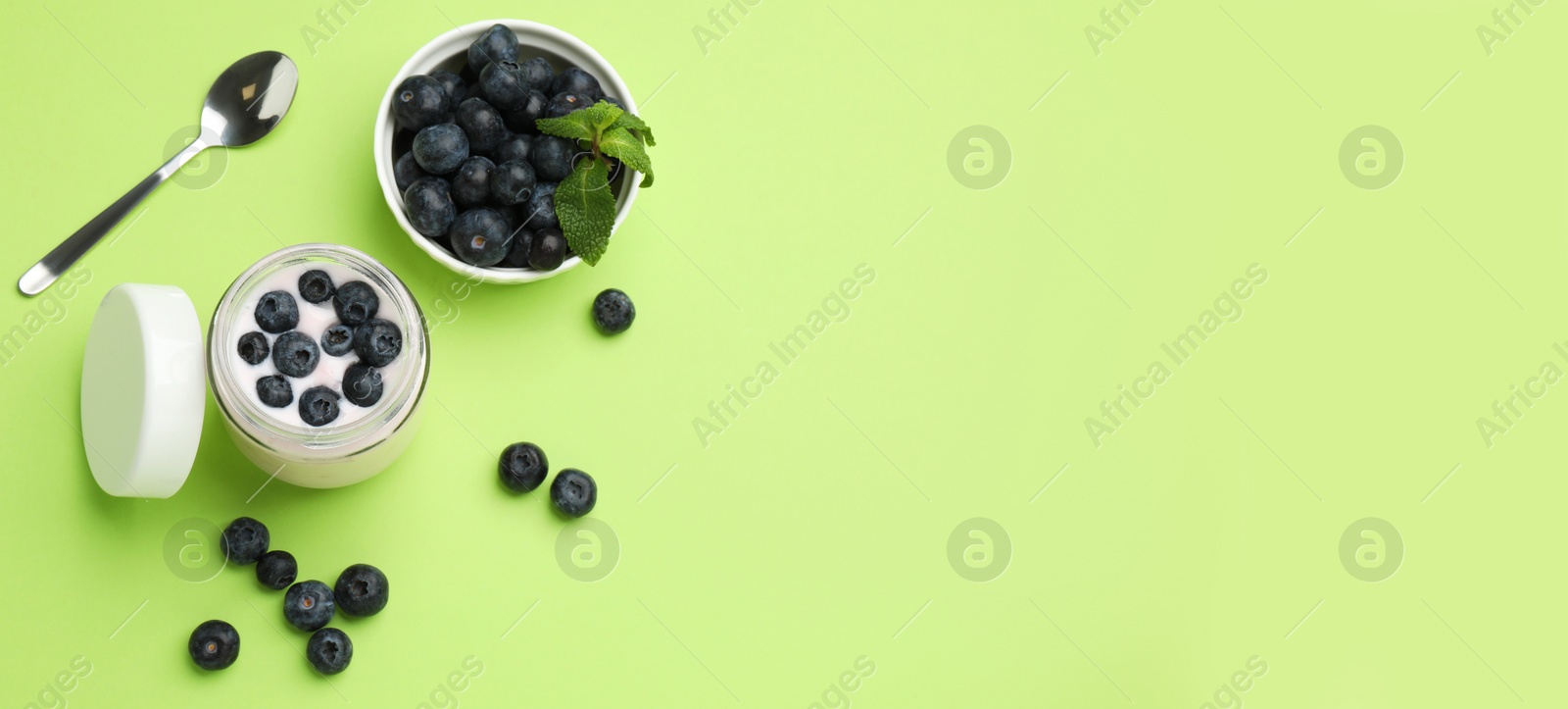 Image of Jar of fresh yogurt with blueberries and spoon on light green background, flat lay with space for text. Banner design