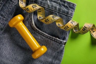 Jeans, dumbbell and measuring tape on green background, flat lay. Weight loss concept