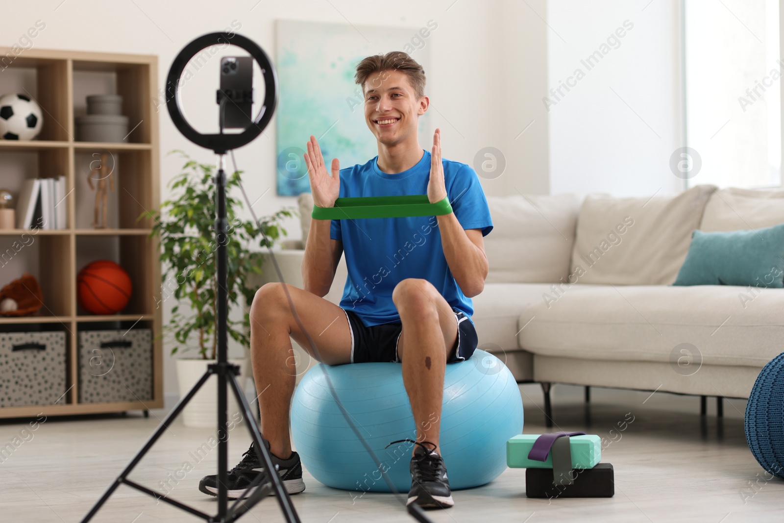 Photo of Smiling sports blogger with resistance band streaming online fitness lesson with smartphone at home