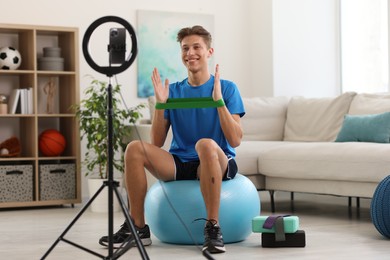 Photo of Smiling sports blogger with resistance band streaming online fitness lesson with smartphone at home