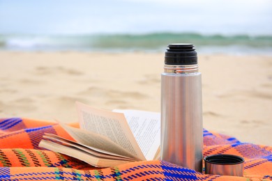 Metallic thermos with hot drink, open book and plaid on sandy beach near sea