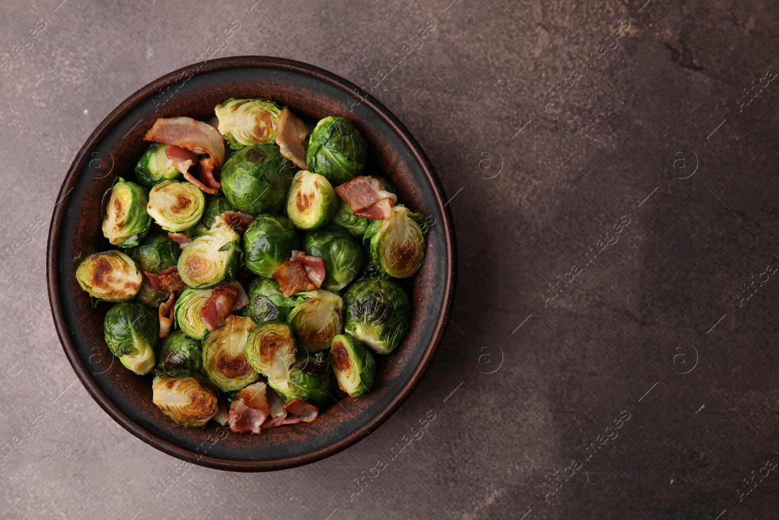 Photo of Delicious roasted Brussels sprouts and bacon in bowl on brown table, top view. Space for text