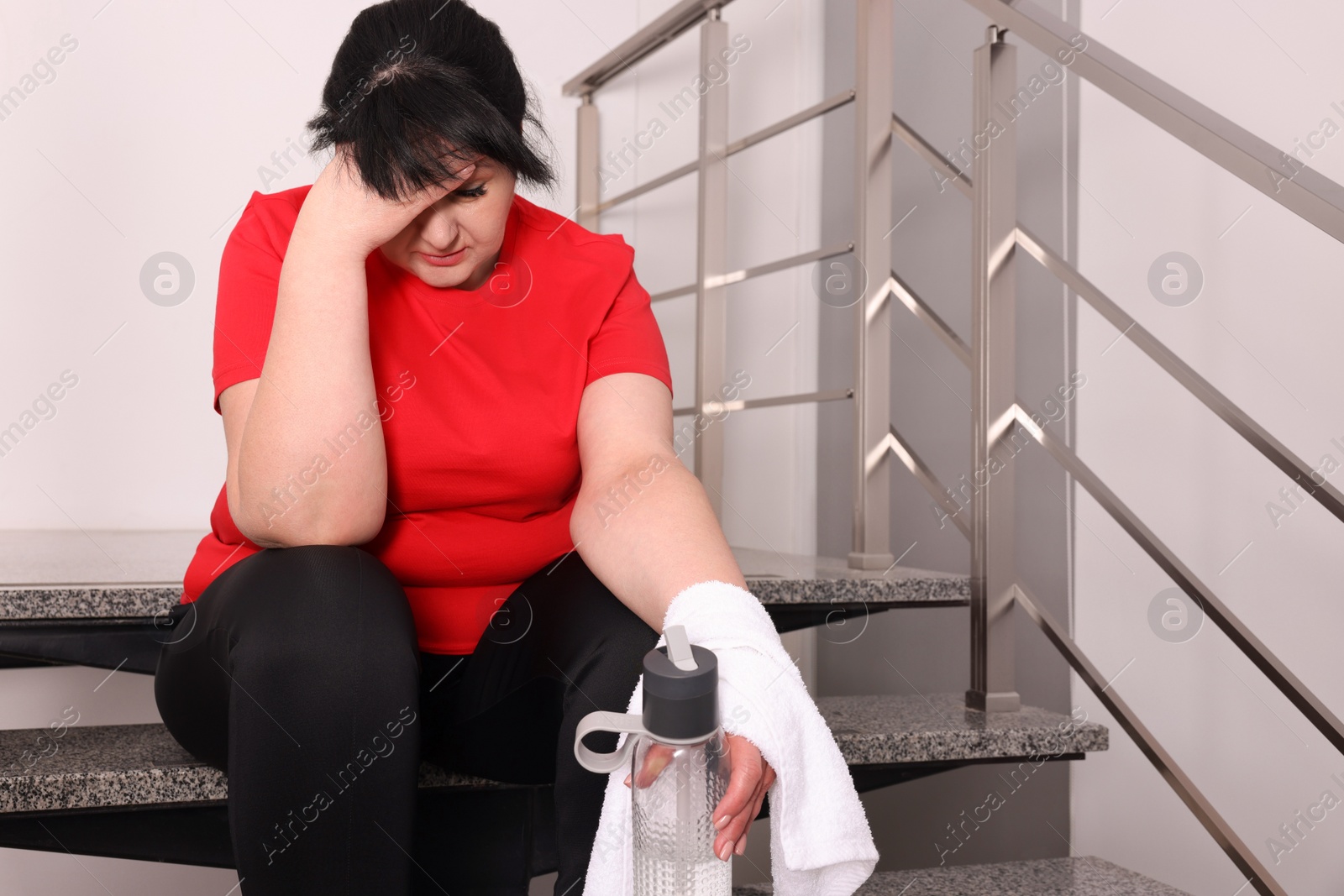 Photo of Tired overweight mature woman with towel and bottle of water sitting on stairs indoors