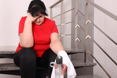Photo of Tired overweight mature woman with towel and bottle of water sitting on stairs indoors
