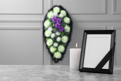 Photo of Photo frame with black ribbon, burning candle on light grey table and wreath of plastic flowers near wall indoors, space for text. Funeral attributes