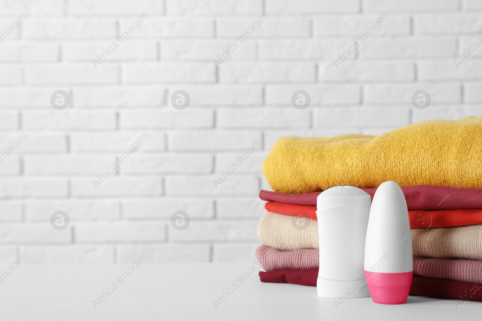 Photo of Deodorants near stack of clean clothes on white table. Space for text