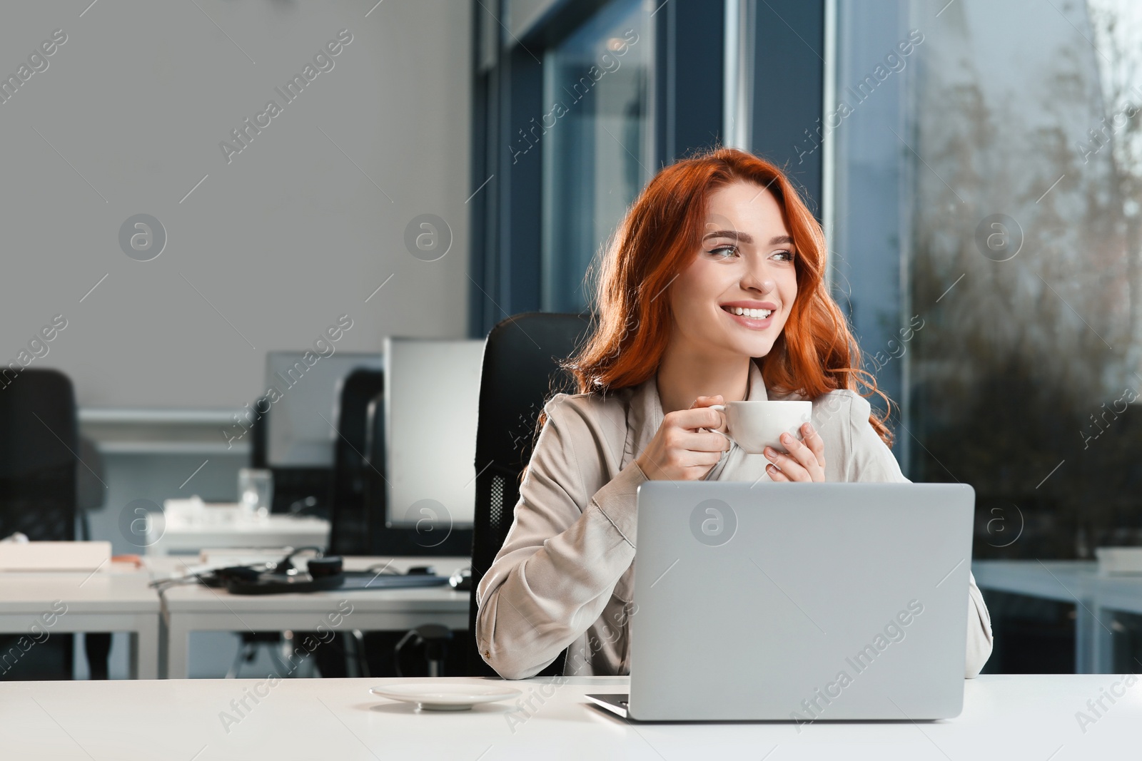 Photo of Happy woman with cup of drink working on laptop at white desk in office