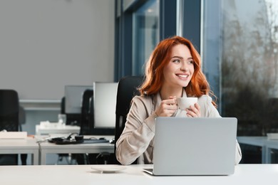Happy woman with cup of drink working on laptop at white desk in office