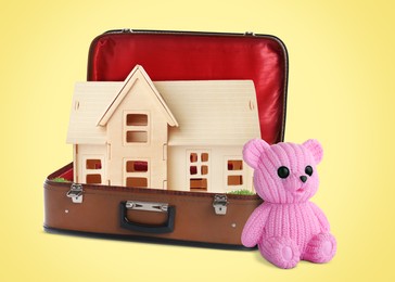 Image of Cute toy bear near retro suitcase with wooden model of house on yellow background