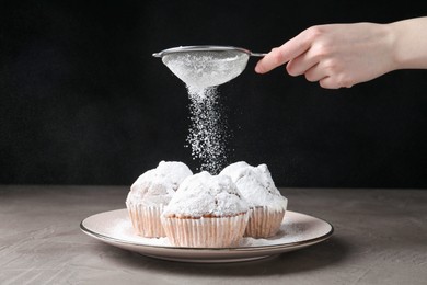 Woman with sieve sprinkling powdered sugar onto muffins at grey textured table, closeup