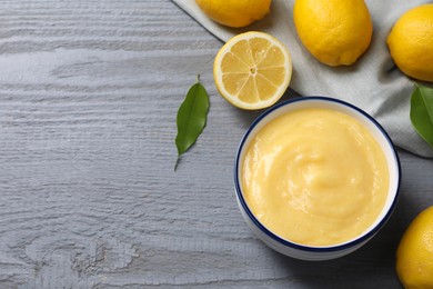 Photo of Delicious lemon curd in bowl, fresh citrus fruits and green leaves on grey wooden table, above view. Space for text