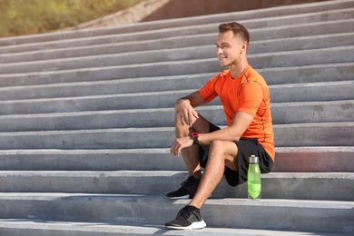 Young sporty man with bottle of water sitting on stone stairs on sunny day