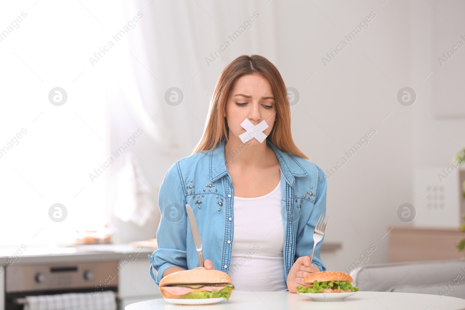 Photo of Sad young woman with taped mouth and burgers at table in kitchen. Healthy diet