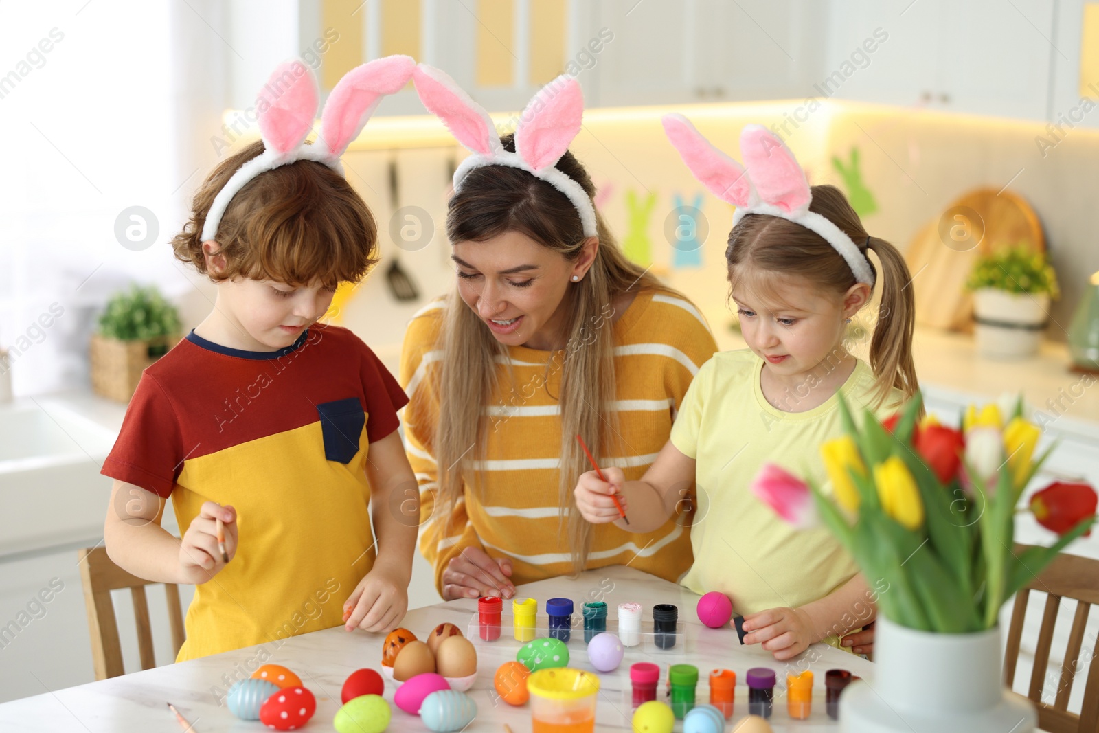 Photo of Easter celebration. Mother and her cute children with bunny ears painting eggs at white marble table in kitchen