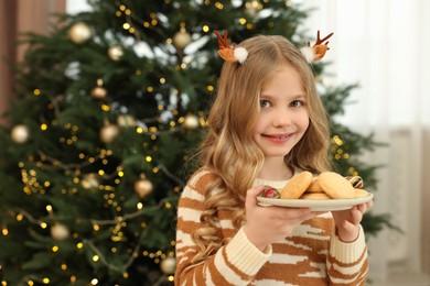 Photo of Portrait of cute little girl in Christmas hair clips holding plate with cookies at home
