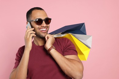 Photo of Happy African American man in sunglasses with shopping bags talking on smartphone against pink background