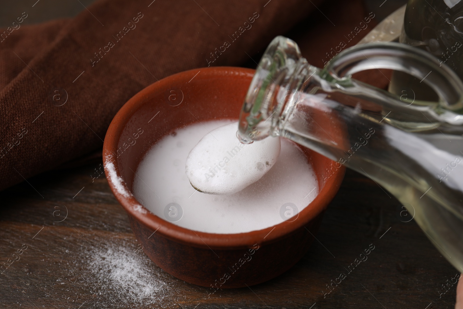 Photo of Pouring vinegar into spoon with baking soda over bowl at wooden table, closeup