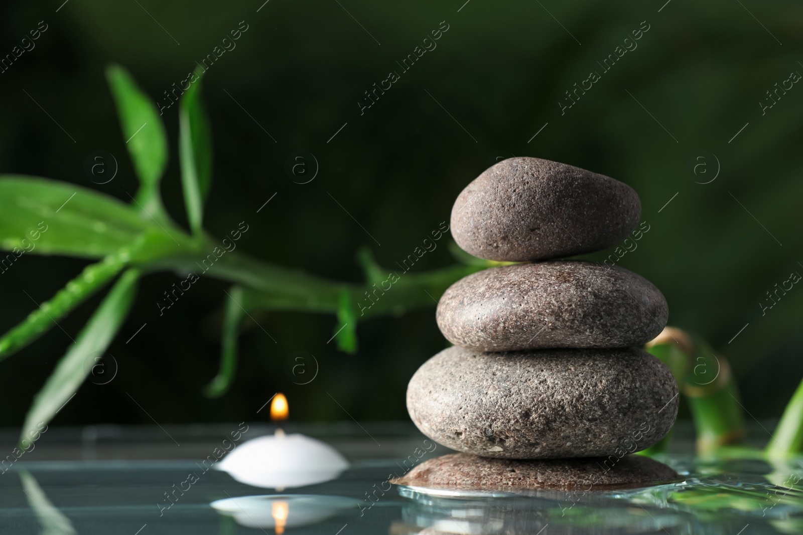 Photo of Stacked stones and burning candle with bamboo stem on water surface against dark background, closeup