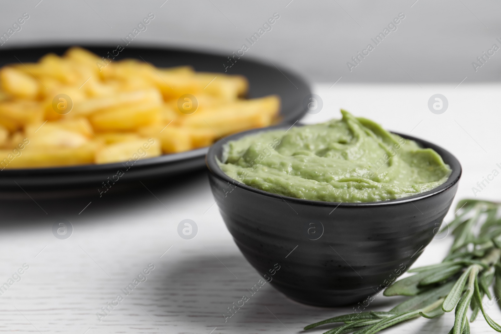 Photo of Dish with avocado dip, rosemary and plate of french fries on white wooden table, closeup