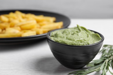 Dish with avocado dip, rosemary and plate of french fries on white wooden table, closeup