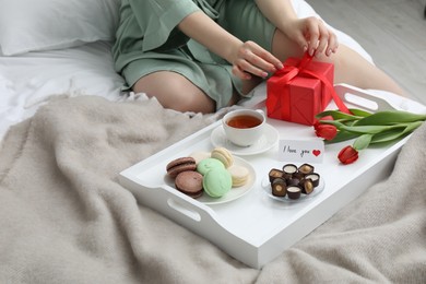 Photo of Tasty breakfast served in bed. Woman with gift box, desserts, tea, flowers and I Love You card at home, closeup