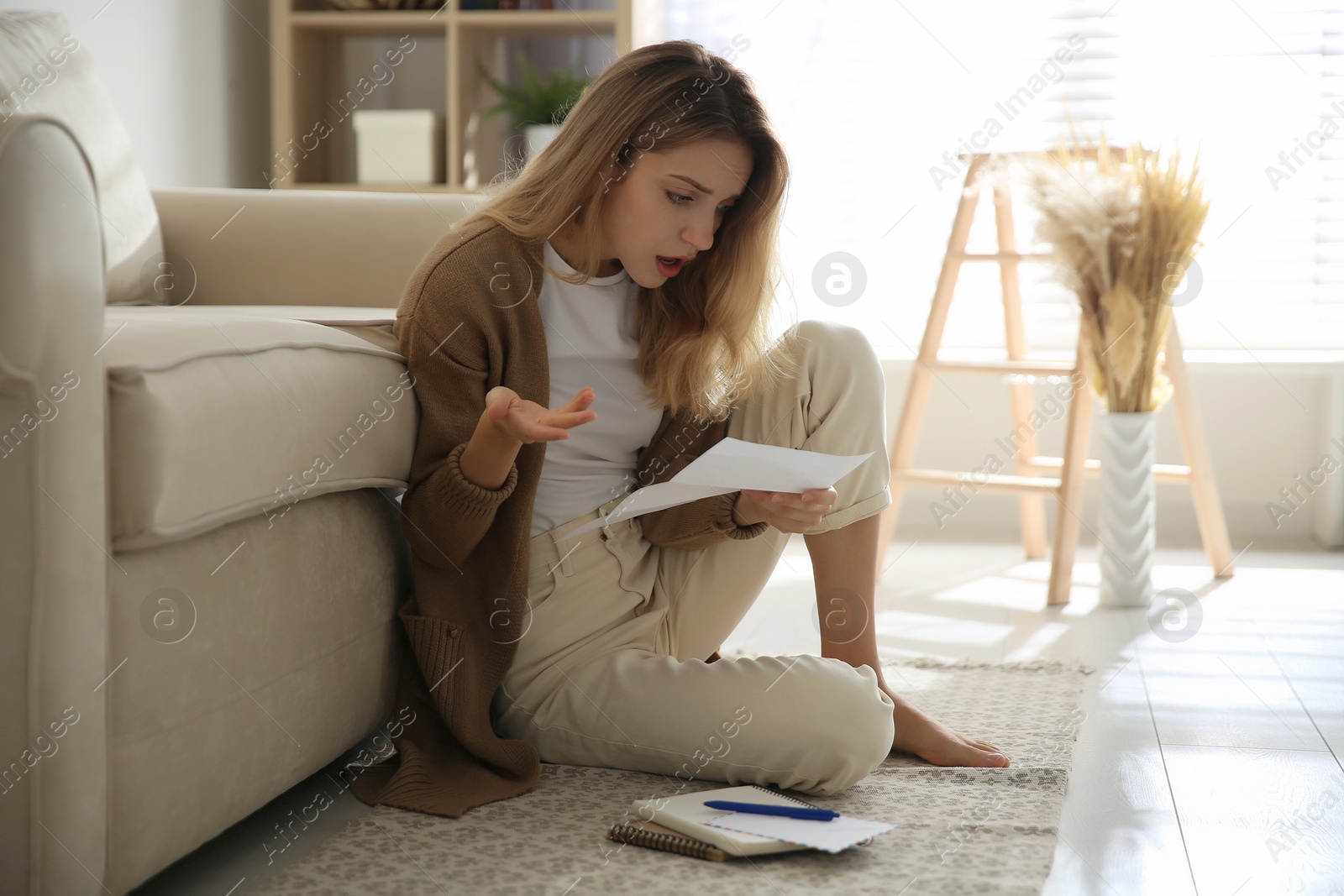 Photo of Shocked woman reading letter while sitting on floor near sofa at home