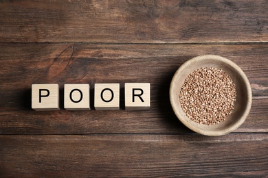 Photo of Bowl of buckwheat and cubes with word POOR on wooden background, top view