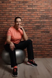Happy overweight woman sitting on fitness ball near brick wall indoors