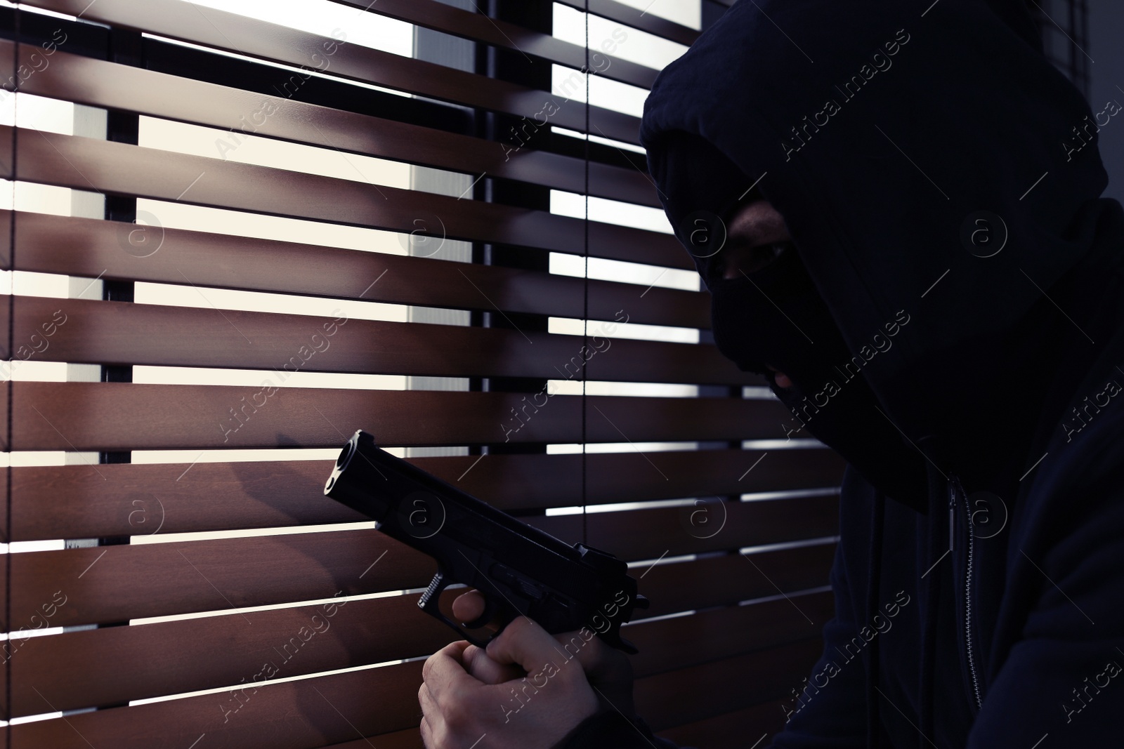 Photo of Masked man with gun spying through window blinds indoors. Dangerous criminal