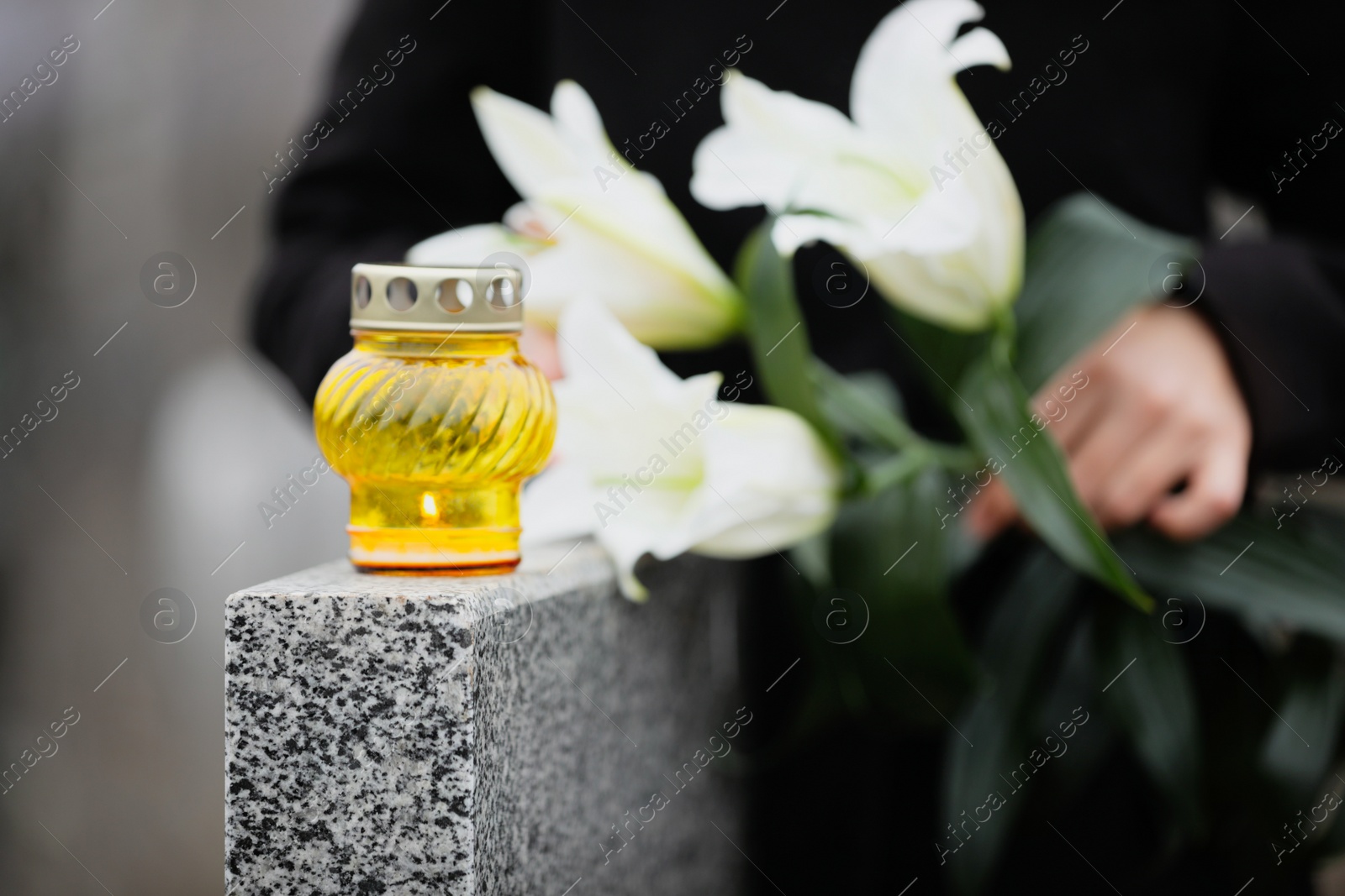 Photo of Woman holding white lilies near grey granite tombstone outdoors, focus on candle. Funeral ceremony