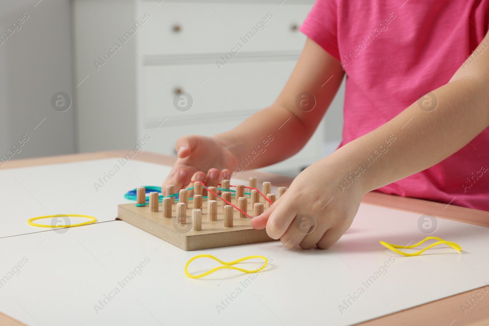 Photo of Motor skills development. Girl playing with geoboard and rubber bands at white table indoors, closeup